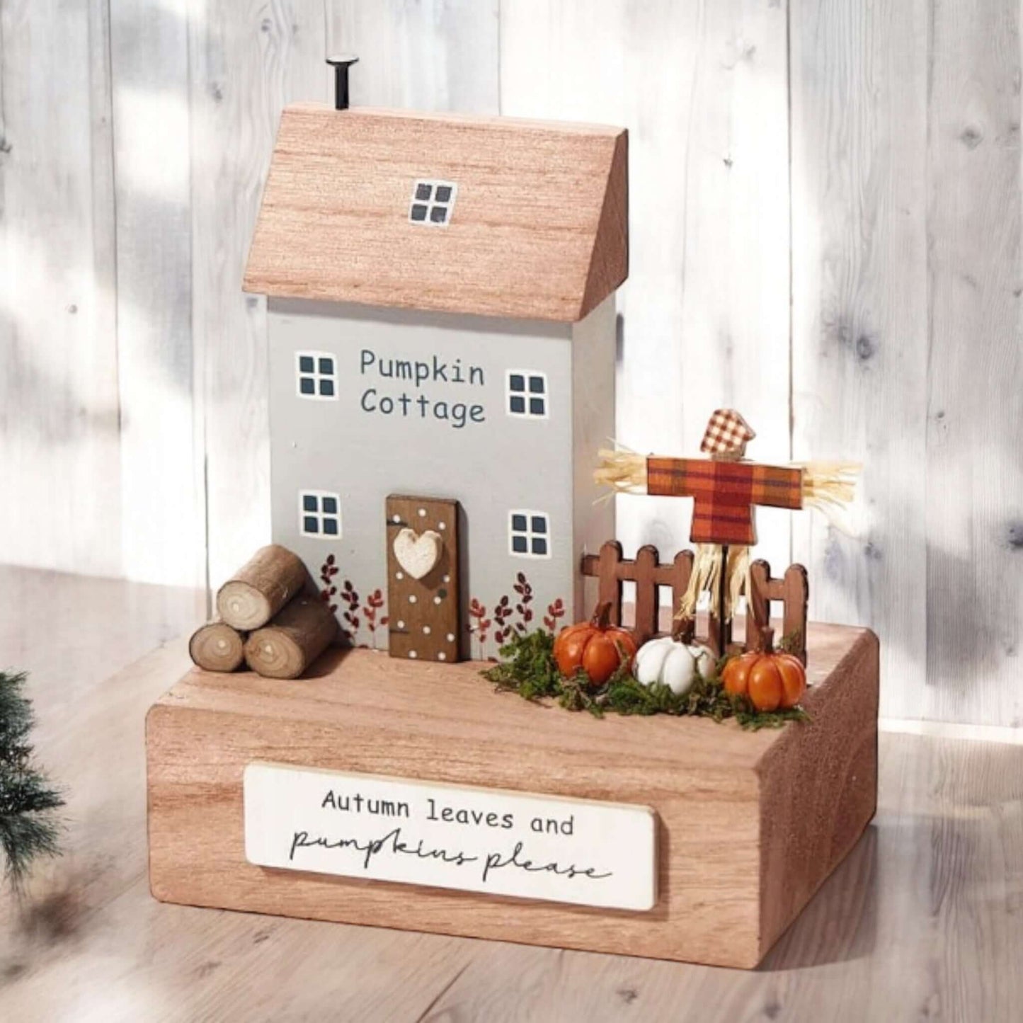 Small grey and natural coloured wooden house named Pumpkin Cottage, featuring a scarecrow alongside a picket fence with pumpkins nearby and a plaque with the words 'Autumn leaves and pumpkins please' on the front. From the Autumn 2024 gift collection at Wickerhearts.