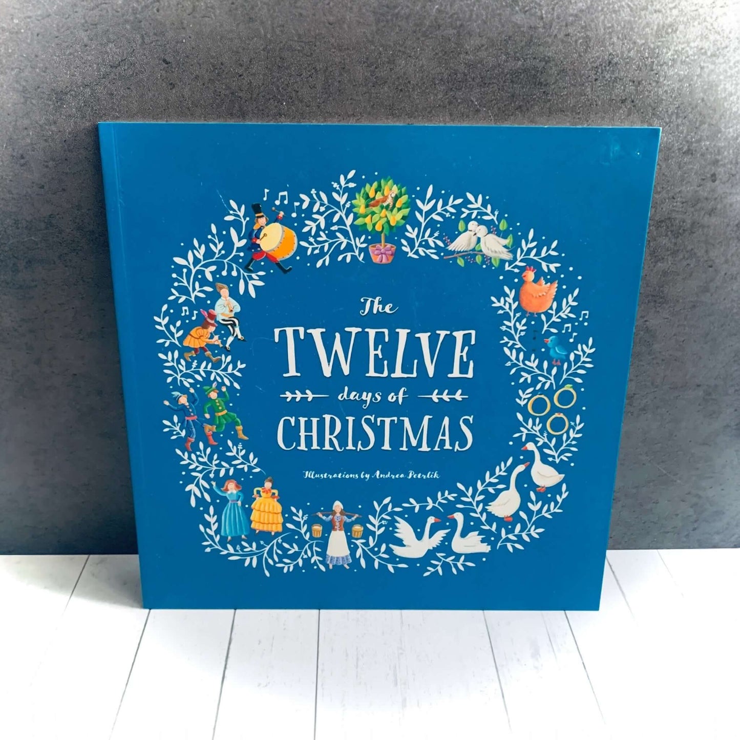 Front cover of the illustrated children's book The twelve days of Christmas, on blue background. Features images from the twelve days of the story. From Wickerhearts Christmas gift collection.