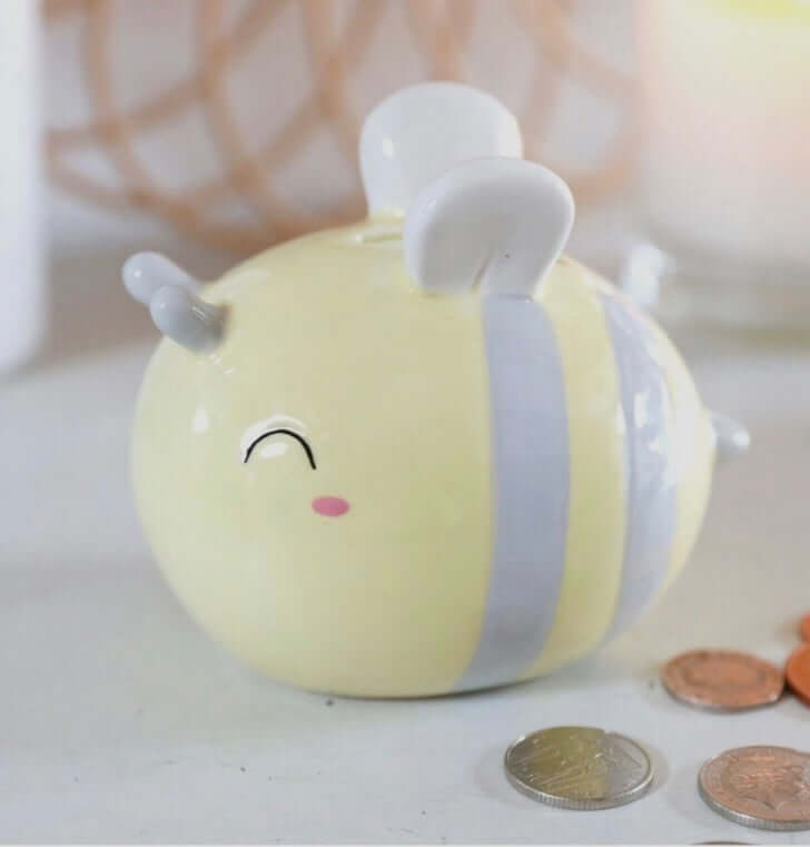 Side view of pale yellow and grey striped bee money box, with pennies by the side of it. From Wickerhearts gift collection.