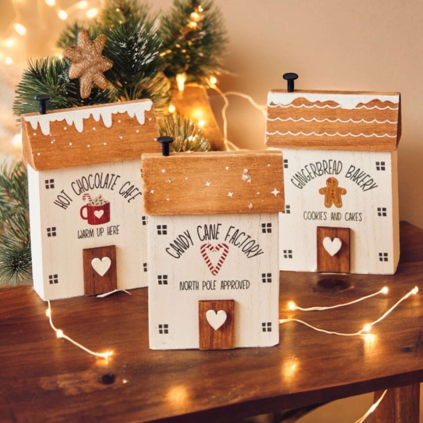 3 little wooden houses with Christmas themes of Candy Cane Factory, Hot Chocolate Cafe or Gingerbread Bakery on a wooden counter top surrounded with Christmas lighting. From the Christmas gift collection at Wickerhearts.