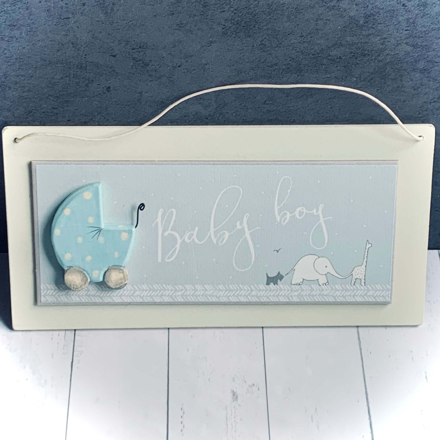 Light wood horizontal sign with blue centre and cream background. Wooden decor pram on left side and picture of giraffe, elephant and dog linked together bottom right. Words are 'Baby Boy'. From Wickerhearts East of India gift collection.