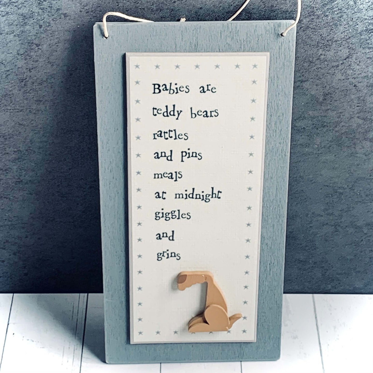 Long grey, light wood hanging picture with brown dog at the bottom and the words 'Babies are teddy bears rattles and pins meals at midnight giggles and grins'. From Wickerhearts East of India gift collection.