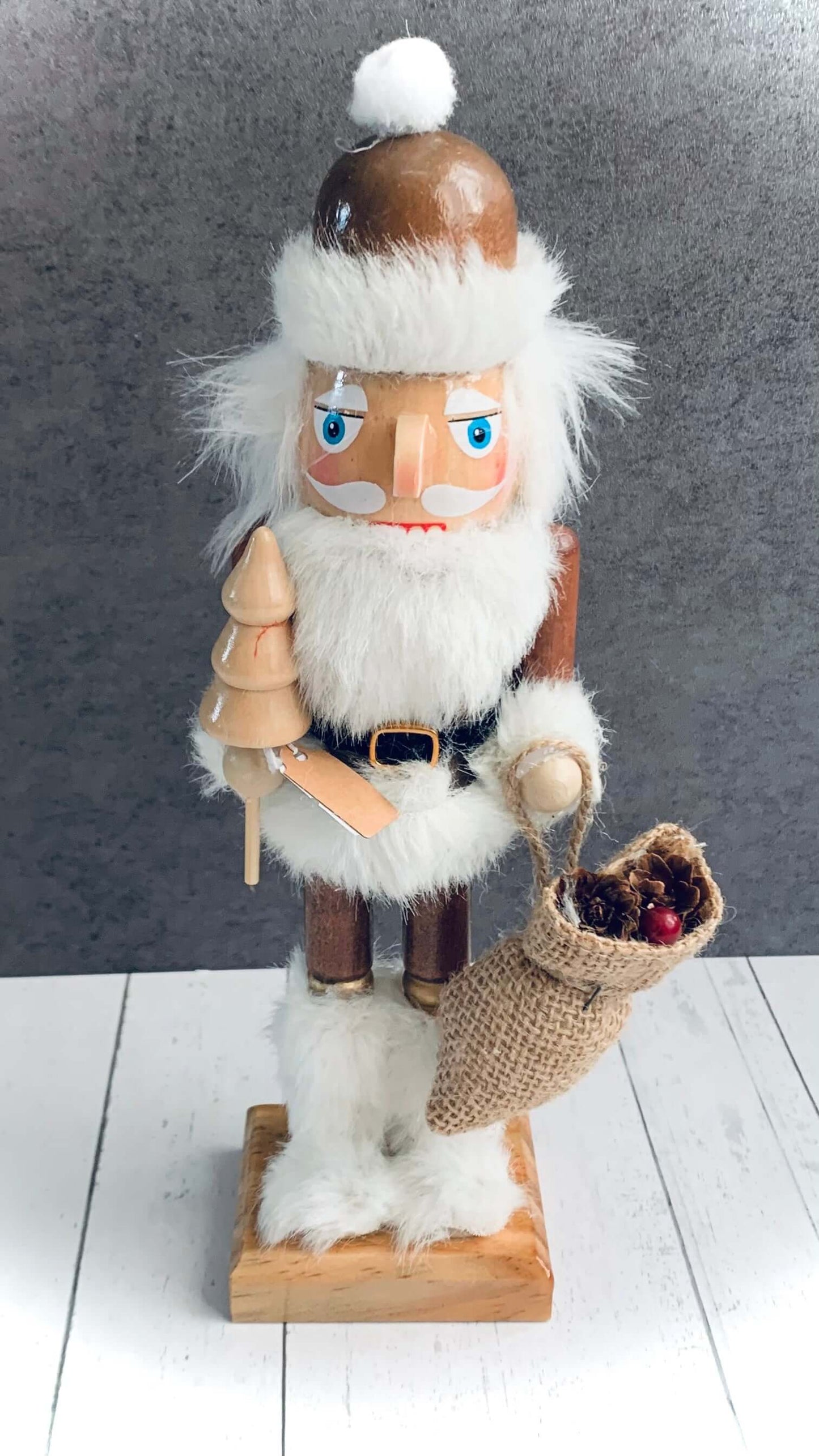 Image of Nordic Santa Nutcracker decoraton, with white beard and hair and white furry boots carrying a small wooden tree and a bag of goodies. From Wickerhearts Christmas gift collection.