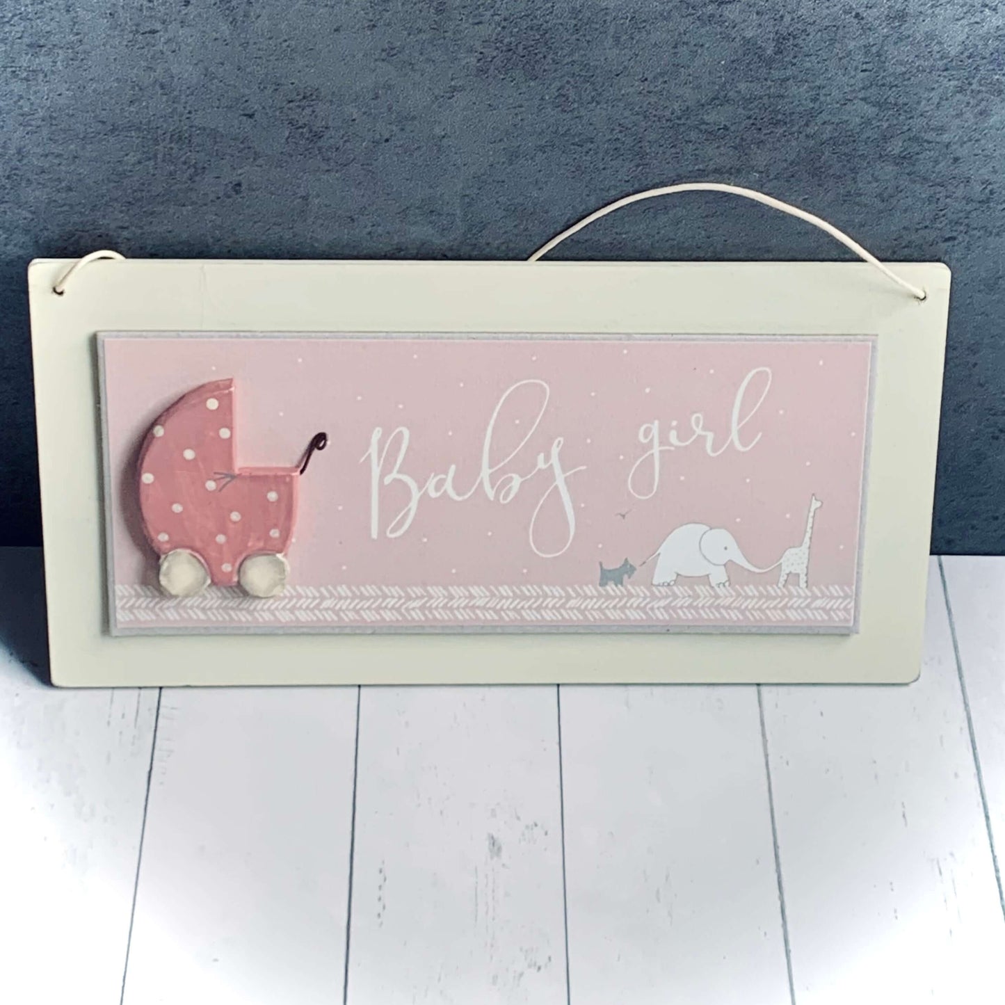 Light wood, horizontal hanging sign in cream and pink for a baby girl. Wooden pink pram on left side and giraffe, elephant and dog linked together bottom right. Words are 'Baby Girl'. From Wickerhearts East of India gift collection.