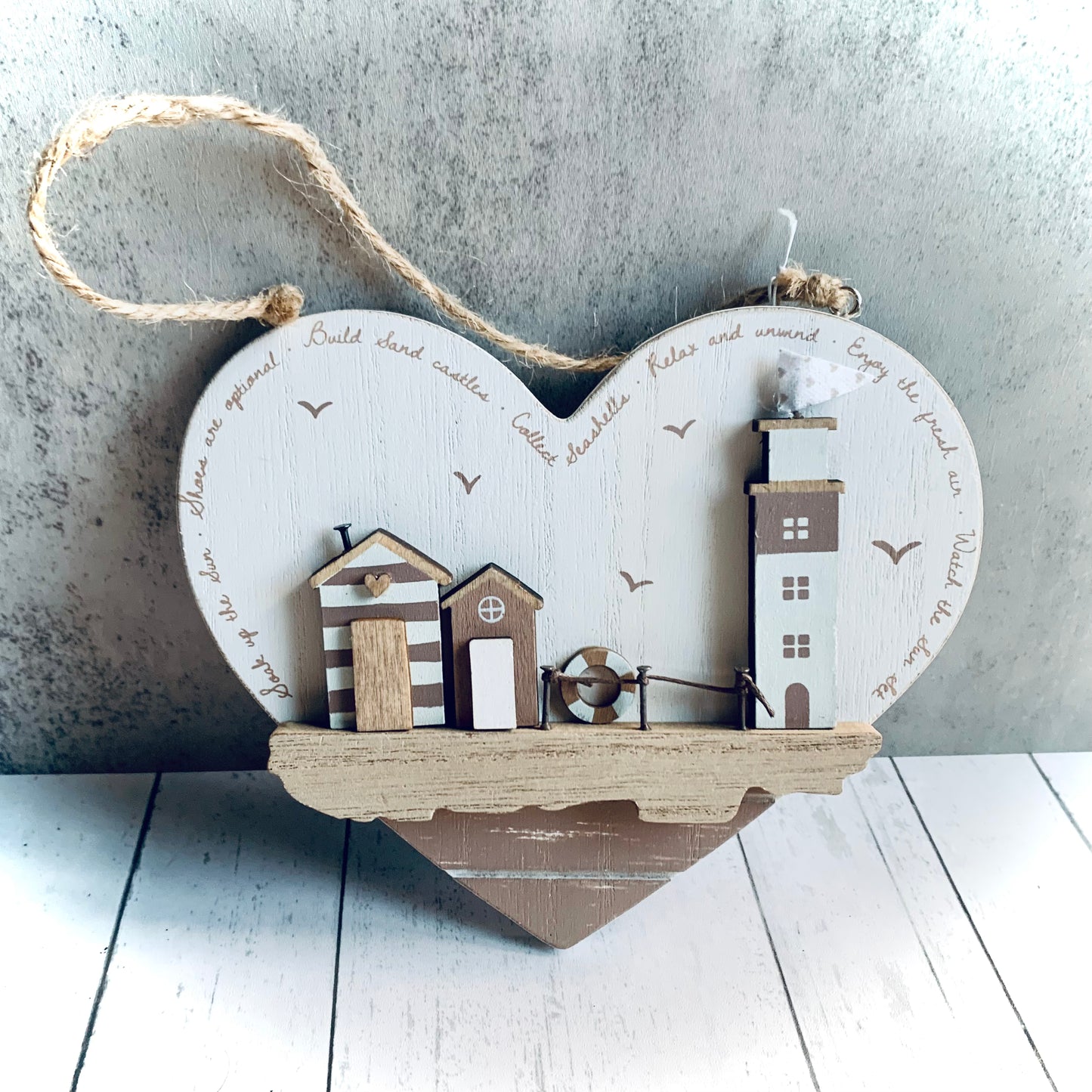 Beach themed wooden hanging heart, with jute hanger and features a lighthouse and beach huts on a quayside in neutral white and beige colours. From the Spring 2024 collection at Wickerhearts.