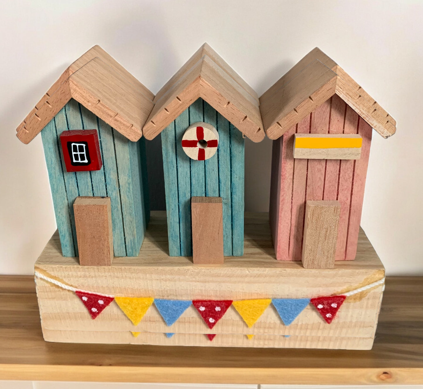 Beach Hut Ornament typically for bathroom decor, consists of 3 wooden beach huts in blue and pink on a light wooden base with colourful bunting. From the Gift collection at Wickerhearts.