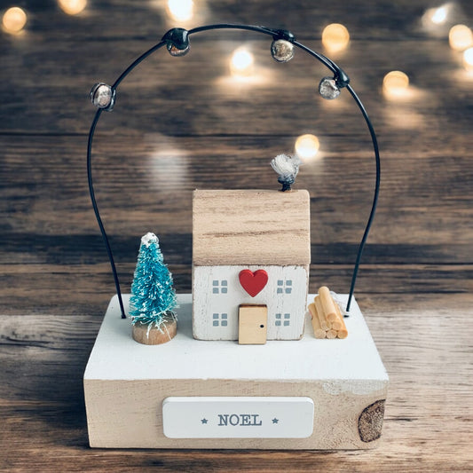 Small wooden house with heart on wooden block, wire above with small bells on. The word 'Noel' on front of block. On a rustic tabletop with subtle lights behind. From Wickerhearts Christmas gift collection.