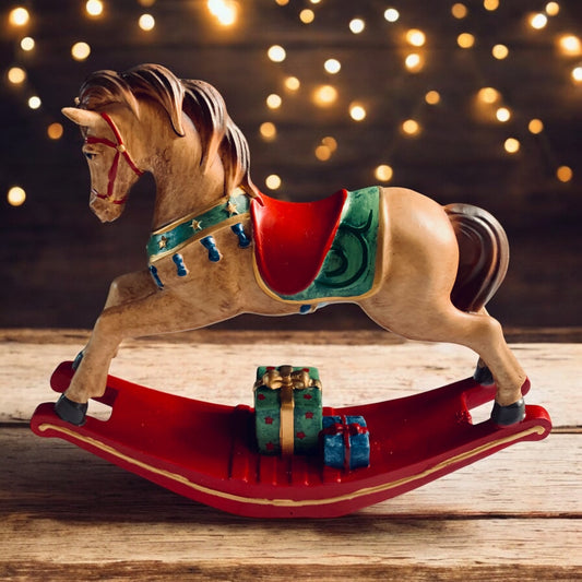 Elegant light brown rocking horse decoration with red saddle and gift parcels underneath. On rustic tabletop with subtle background lights. From Wickerhearts Christmas gift collection.