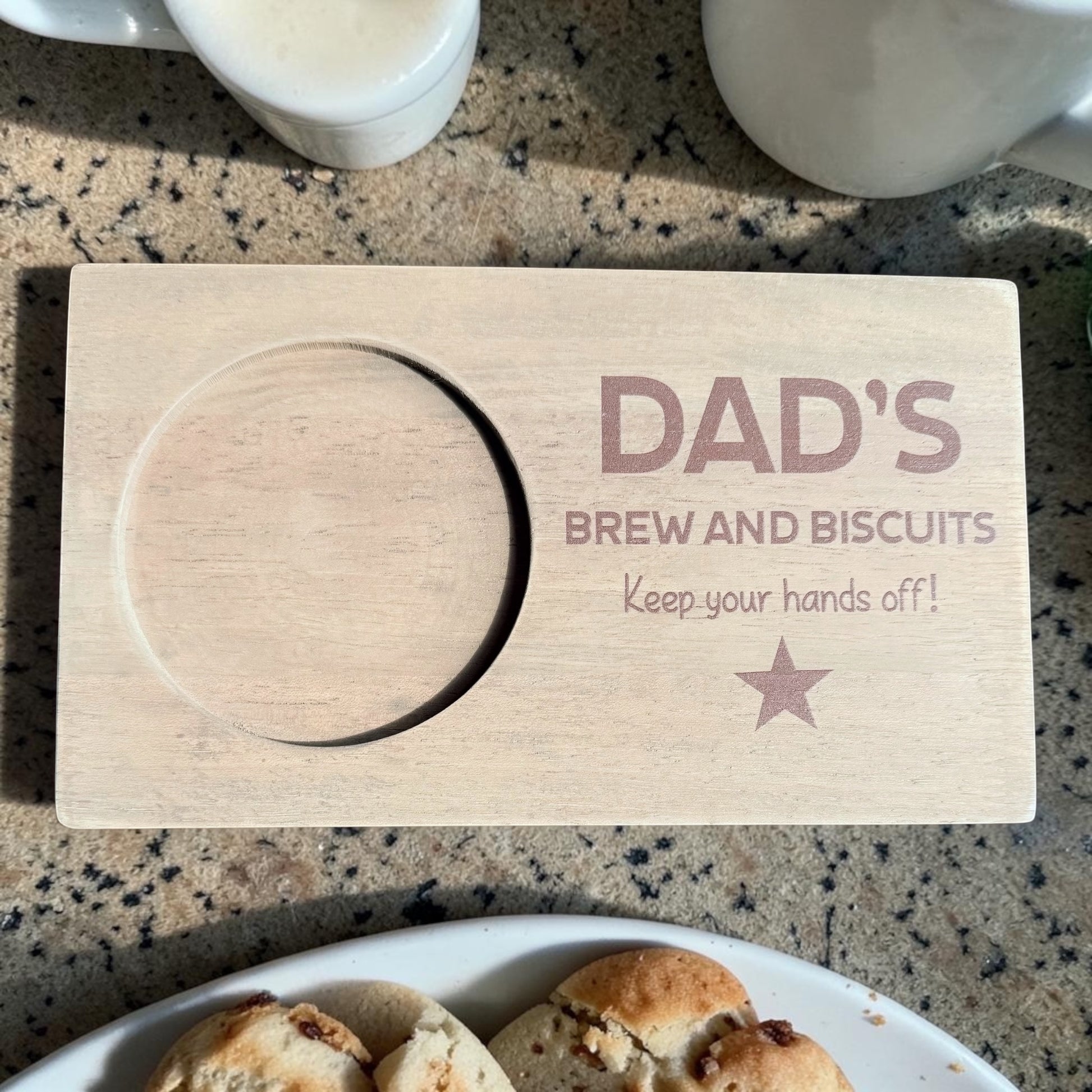 Wooden coaster with 'DAD'S BREW AND BISCUITS Keep your hands off!' text on a countertop with a plate of cookies and cups.