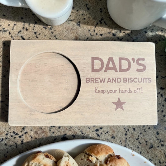 Wooden coaster with 'DAD'S BREW AND BISCUITS Keep your hands off!' text on a countertop with a plate of cookies and cups.