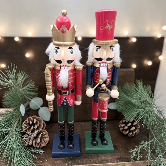 Two decorative Christmas nutcracker figurines standing on a wooden surface, surrounded by pine branches, eucalyptus leaves, and pinecones. One nutcracker wears a red coat and green pants with a gold crown, and the other wears a red coat with blue pants and a tall red hat.